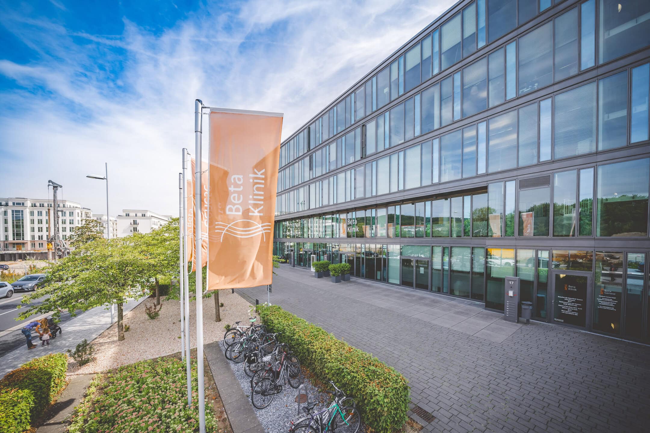 Eine orangene Beta Klinik Fahne im Vordergrund des Klinik Haupteingangs. Ein modernes, aus Glas bestehendes, Gebäude.