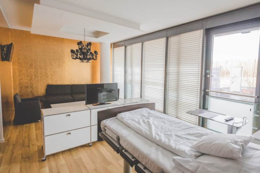 Modernes Patientenzimmer mit großer Fensterfront, Fernseher und Sitzlounge
