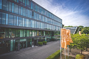 Beta Klinik: Große Glasfront mit Haupteingang und orangen Flaggen vor dem Gebäude.