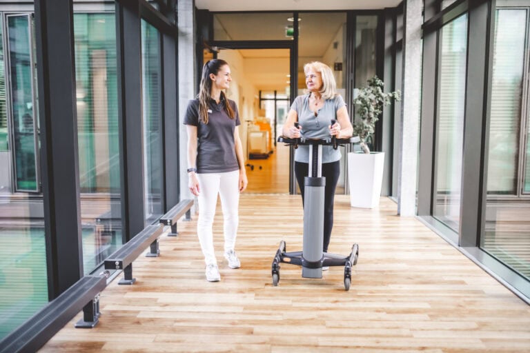 Patientin mit Gehunterstützung wird von Pflegerin durch Klinikgebäude begleitet.