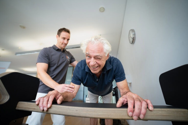 Patient lächelt in die Kamera, während er sich auf einem Five-Parcours Gerät abstützt und vom Physiotherapeut begleitet wird.